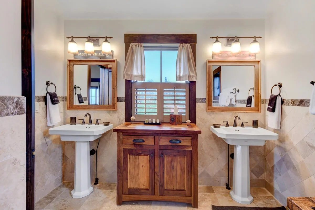 A spacious bathroom with double sink vanities, large mirrors, a wooden cabinet, and a window filling the space with natural light. The bathroom exudes calm with its clean lines and serene atmosphere, ideal for self-care and relaxation.