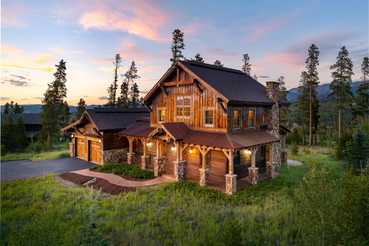 The image showcases a luxurious wooden single-family home framed by stone accents, with a spacious wraparound porch overlooking lush greenery. The warm glow of the sunset sky adds a cozy, inviting touch, hinting at memorable evenings spent outdoors in the serene Colorado landscape.