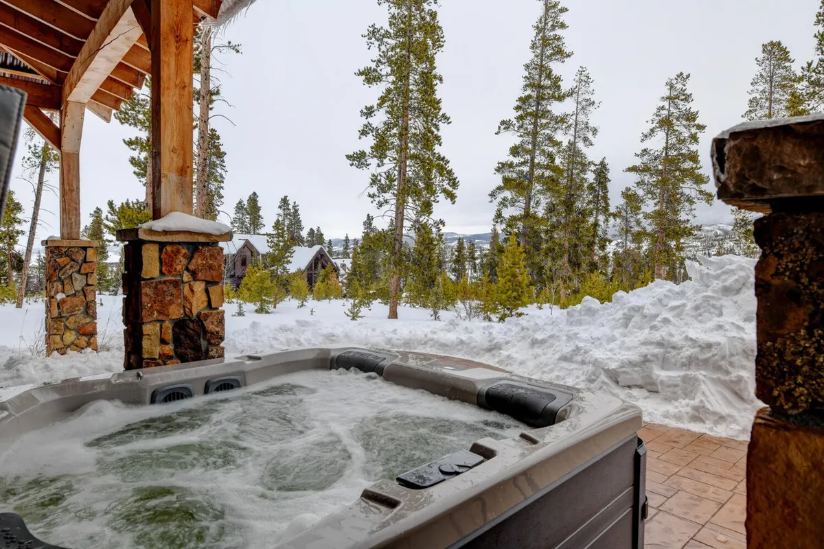 An inviting outdoor hot tub set amid a snow-covered landscape and tall pine trees, with stone pillars framing the scene. The hot tub is steaming gently, suggesting warmth and comfort, while the open sky above hints at opportunities for stargazing. The surrounding mountains and wintery environment create a tranquil and luxurious atmosphere.