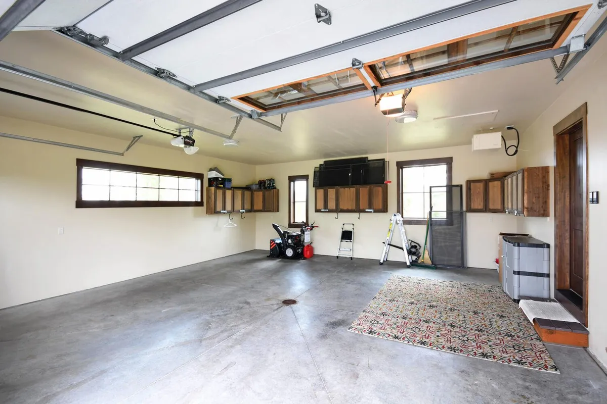 A functional interior view of a garage with a visible garage door, storage cabinets, a workbench, and an area rug. The space is organized and inviting, suited for storing vehicles, bikes, and gear. The heated, spacious environment enables easy transitions between outdoor and indoor activities throughout the seasons.