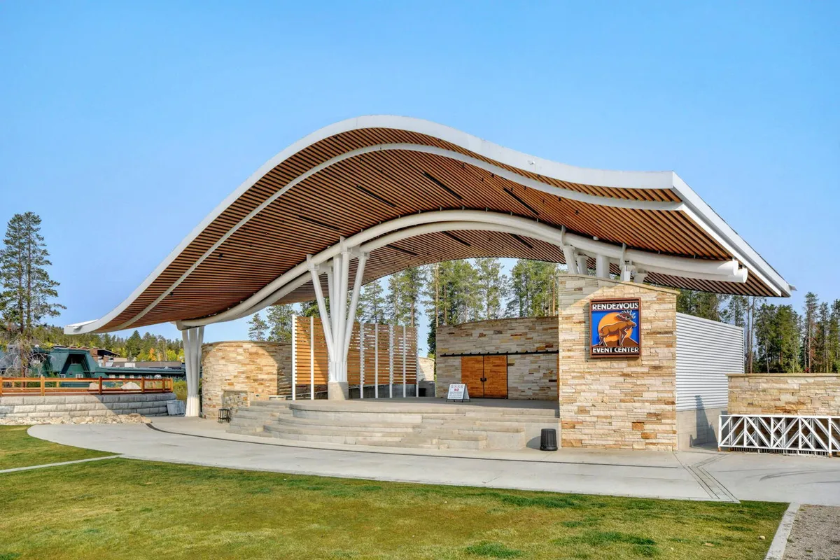 A vibrant community space with a unique roof design, stone walls, and an open stage area set up for concerts or outdoor events. The scene evokes feelings of festivity, with plenty of room for families to gather and enjoy entertainment together.