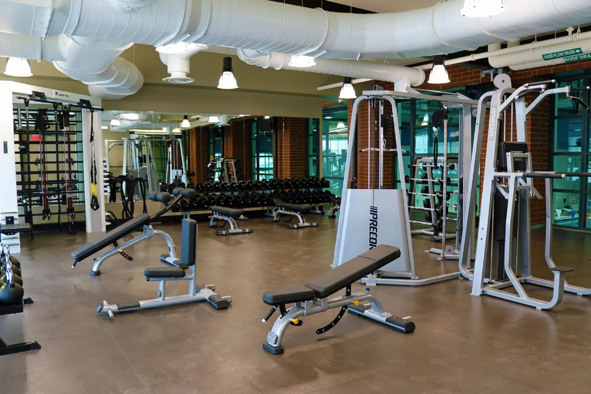 This interior photo showcases a modern fitness space with weightlifting equipment, exercise benches, mirrored walls, and a neatly organized dumbbell rack, reflecting a highly functional gym environment.