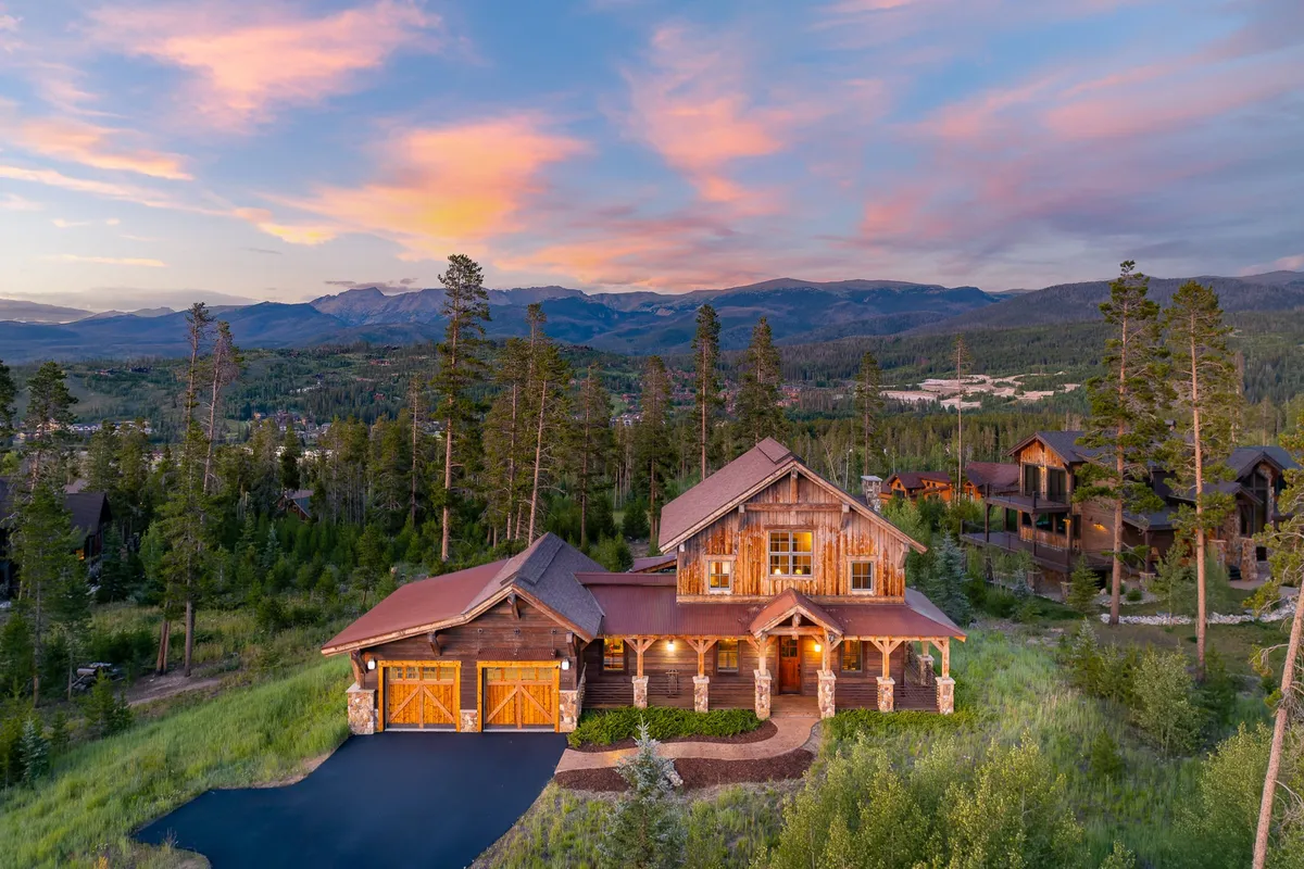 This exterior image captures the rustic wooden façade of the luxury home, set against a vibrant sunset sky and surrounded by towering pine trees. The inviting porch and two-car garage highlight the home's grandeur and exclusivity, while the tranquil natural environment perfectly frames the home within the prestigious Leland Creek neighborhood.