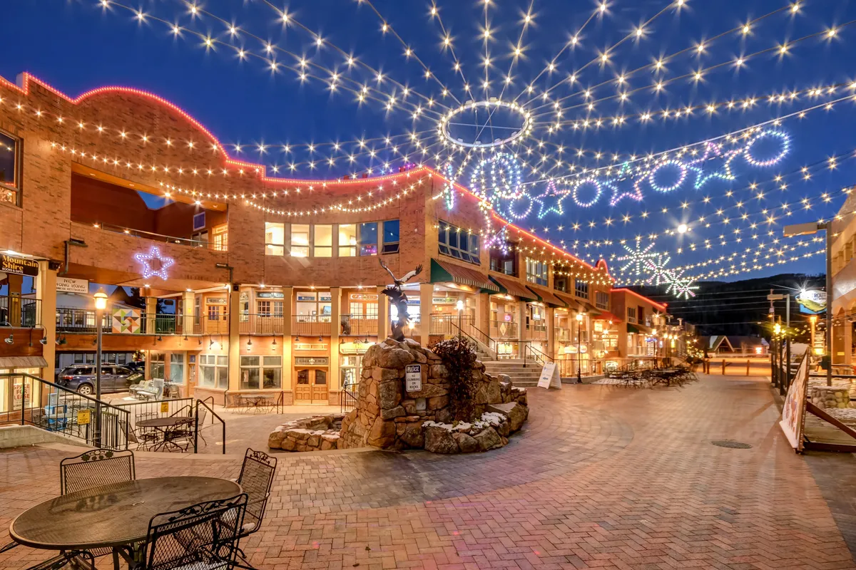A charming pedestrian-friendly area with decorative string lights, brick walkways, outdoor seating, a sculpture fountain, and boutique storefronts. The scene suggests an inviting environment for dining, shopping, and seasonal events.